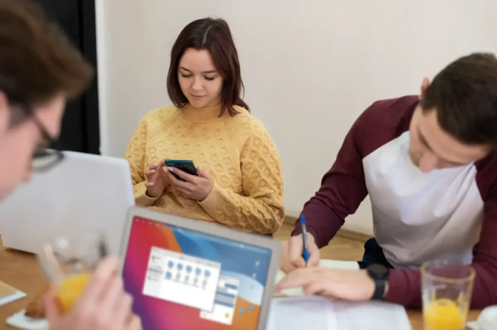 Personas trabajan sobre una mesa. Una mujer sostiene un celular.