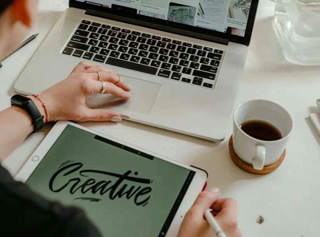 Persona trabajando sobre una mesa, en ella se encuentra una tablet con la palabra "creative", un café y un computador.