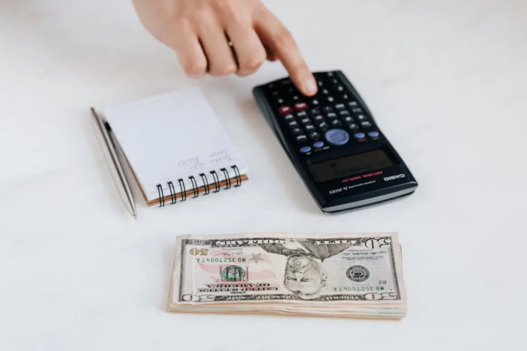 Una mano aprieta un botón de la calculadora que está sobre una mesa blanca junto a unos billetes y una libreta.